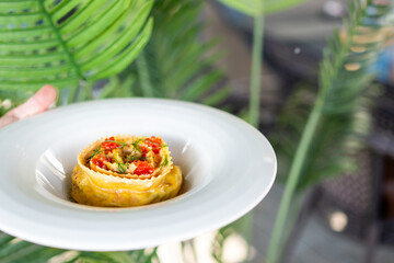 Elegant gourmet dish with rolled pasta stuffed with filling, garnished with red caviar and herbs, served on a white plate against lush green foliage.