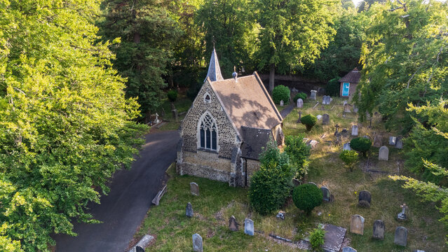 Aerial view of a historic church nestled among vibrant green trees and aged gravestones, its stone facade bathed in soft light, Guildford, England, United Kingdom.