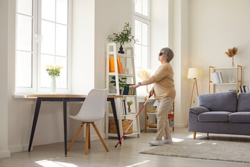 Old woman having problems with eyesight, being blind, walking with cane in living room, being alone. Blindness, disability and vision problems concept. Blind mature female at home. 