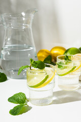 Glass cups and a jug of clean, clear drinking lemon and lime water on a white table. Sunny day and sharp shadows from glass of lemonade