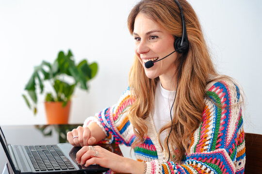 Smiling young woman in vibrant crochet sweater sits at a desk wearing a headset, typing on a laptop while engaged in a video call or online meeting, representing remote work and customer support. 