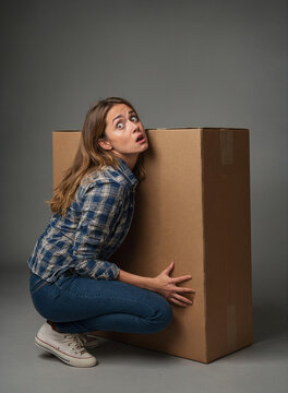Young woman with a frightened and surprised look on her face unsuccessfully tries to lift a huge heavy box.