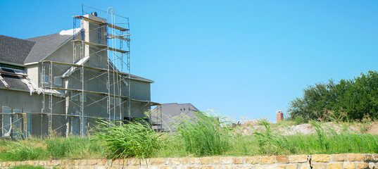 Panorama view Dallas house under construction with chimney framed in scaffolding. Low stone wall separates property from street, highlights woodwork, roof installation, active development zone