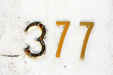 Close-up of weathered metallic house number 377 on a white textured wall. Golden numerals show age...