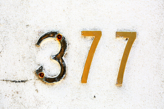 Close-up of weathered metallic house number 377 on a white textured wall. Golden numerals show age and rust, providing a vintage, urban look ideal for architecture or design-related themes - Powered by Adobe