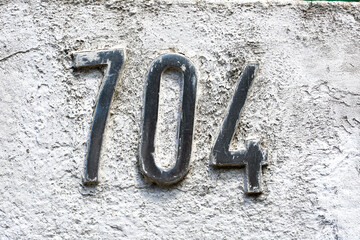 Close-up of metallic house number "704" mounted on a rough, aged concrete wall. The silver numbers show signs of weathering, creating a vintage, textured look with urban and industrial tones
