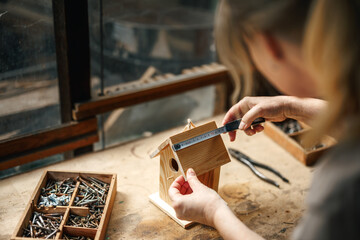 Female artisan measuring wood to make homemade birdhouse, surrounded by vintage tools and DIY...