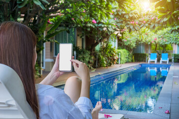 a woman using finger for touch on phone mockup with blank white screen with copy space, relaxing by the poolside on vacation day at hotel resort.