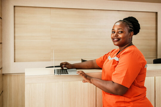 Smiling female nurse in bright orange scrubs standing at hospital reception desk with a laptop, representing healthcare staff, patient service, and hospital administration in a clinical setting.