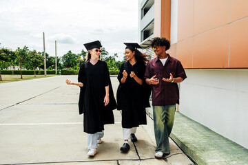 Three happy young graduates walk together outdoors, smiling and chatting after their ceremony. They...