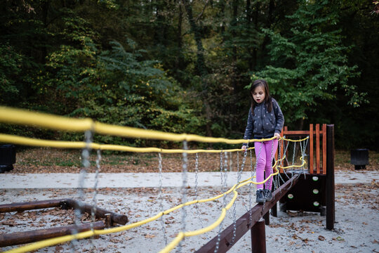 Young girl carefully walking on rope bridge in playground surrounded by trees