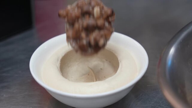 Close-up of spoon adding warm fava beans to creamy hummus, highlighting authentic Middle Eastern cuisine, perfect for cooking channels and food blogs.
