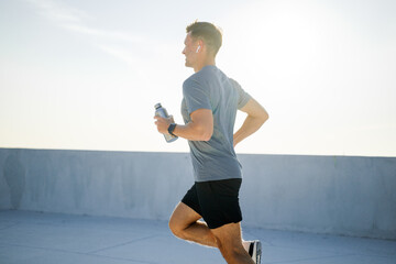 Man running on rooftop during sunrise while holding a water bottle and wearing wireless headphones