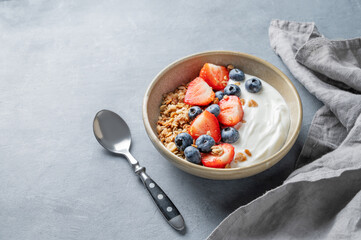 Natural yogurt with granola, blueberry and strawberry in a bowl on a blue background with spoon and napkin.