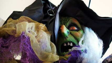Green scary witch mask with crooked teeth and orange eyes, wearing black hat and colorful rags, hanging during Halloween celebration, creating spooky atmosphere