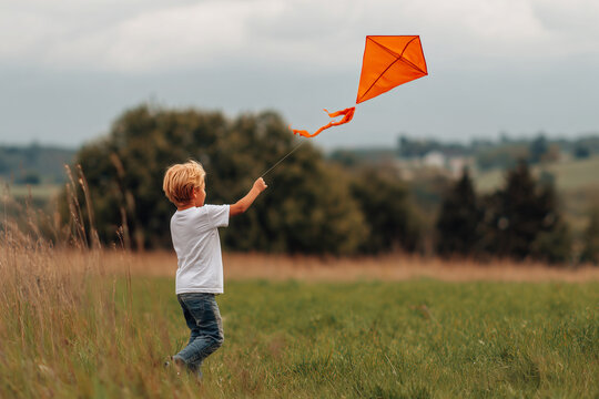 boy flying orange kite outdoors in green field under cloudy sky during daytime - Powered by Adobe