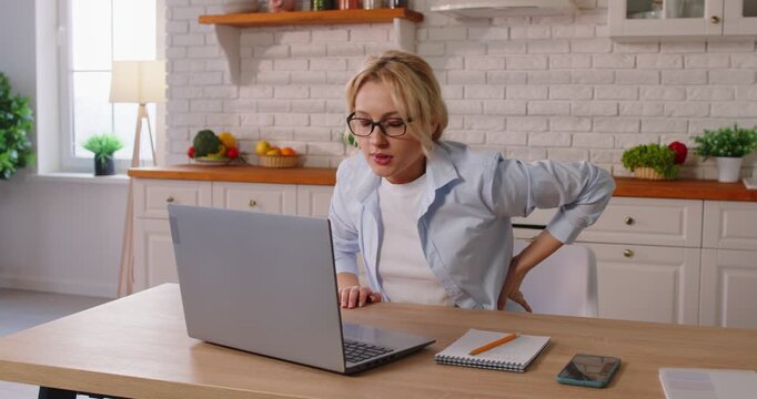Tired woman working online with a laptop at the kitchen table, suffering from back ache and burnout at home. Challenges of remote work, health issues, pain, and the need for self-care.