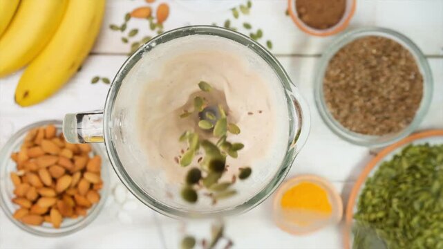 Top view of pumpkin or squash seeds falling into white smoothie blending in a mixer surrounded by ingredients. Vegan plant-based protein shake. No animal fat. Post-workout energy drink.