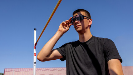 Athlete in sunglasses resting on high jump mat, getting ready to pole vault beneath clear blue sky....
