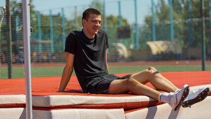 Athlete taking a break on high jump mat at sunny outdoor sports track. Concept of post-effort...
