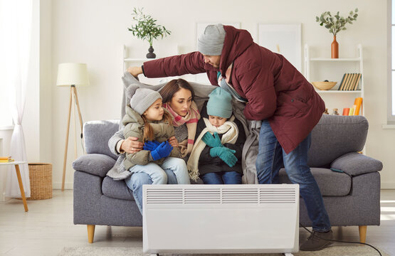 Freezing family trying to warm up near portable electric heater sitting on comfortable sofa wearing jackets and plaid feel very cold in flat during winter time having heating problems at home