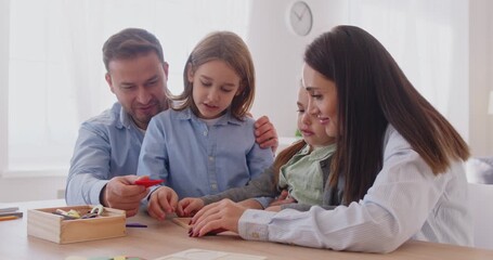 Mother and father playing with wooden geometric figures with little kids at home. Happy family playing logic educational board games with children sitting at desk in living room. 4k video.