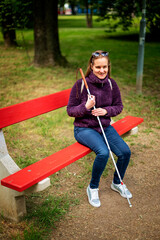A visually impaired woman holding a white cane and sitting on a bench in the park