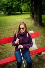 A visually impaired woman holding a white cane and sitting on a bench in the park