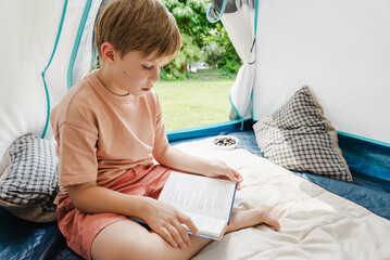 School-age boy sitting in his tent outdoors and reading a book. Summer holidays.
