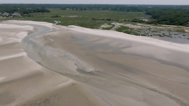 An estuary snakes toward the dunes and low houses, with flowing curves etched in silt and sand below wide skies.