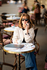 Middle aged woman sitting at sidewalk cafe in the city and using smartphone