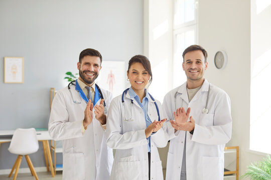 Three cheerful doctors in white coats standing together in medical office, celebrating successful collaboration and achievement. Professional clinic staff team clapping hands and looking at camera.