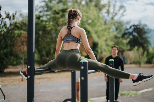 A young woman exercises on outdoor parallel bars in a park setting, performing a split stretch. The scene captures active lifestyle, fitness, and healthy living in a natural environment.