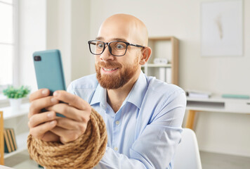 Portrait of a young obsessed business man employee sitting at the desk on workplace in office with his hands tied with rope addicted to mobile phone. Internet and smartphone addiction concept.
