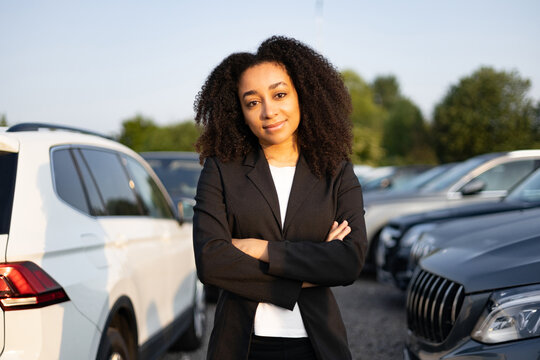 A woman in a business suit stands confidently, arms crossed, at a car dealership, smiling.