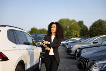 A car salesperson, an African American woman, shows a white SUV to a potential buyer in a car...
