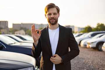 A car salesman gives an ok gesture, standing in front of a car lot, a portrait of a business man in...