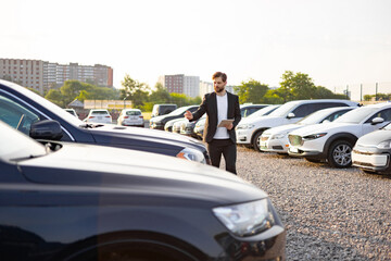 A car salesman in a suit gestures toward a row of cars, holding a tablet, set against a parking lot...