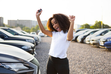 An excited woman celebrates her new car purchase, holding the key high near a car lot with various...