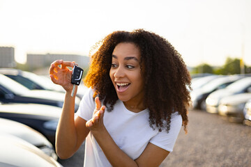 A joyful woman excitedly displays her new car keys, standing in a car dealership parking lot.