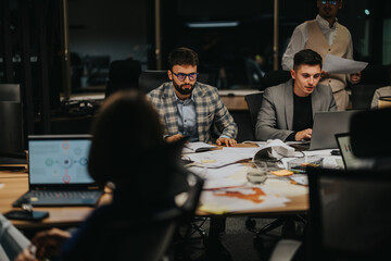 A multicultural team of business professionals collaborates on a project late at night in a modern office setting, with laptops and documents spread across the table.