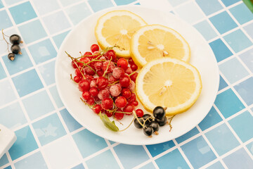 A white plate with three lemon slices, red currants, and black currants, placed on a blue mosaic tile background near a pool. Bright natural sunlight, top-down composition. 