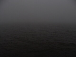Obraz premium Dark, foggy seascape with low visibility, evoking a mysterious and eerie mood over calm, tranquil waters under an overcast sky.