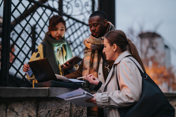 Three diverse businesspeople discussing work outside, engaged in a collaborative session with a laptop and documents, showcasing teamwork and productivity.