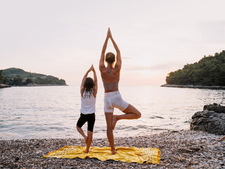 Mother and daughter practicing yoga at seaside at sunset or sunrise.