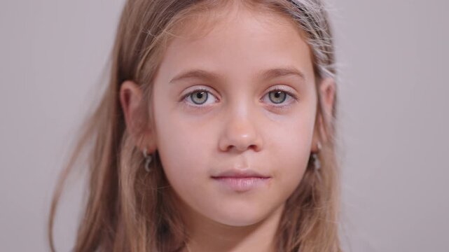 Closeup of young girl with long hair and serious look on face. She is calmly looking at camera indoors with plain background in simple studio lighting
