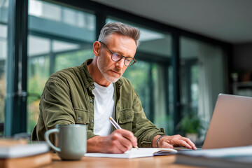 Focused Mature Man Working from Home and Taking Notes at Laptop