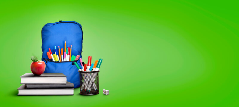 A back to school scene of a pile of three study books to learn from, a red apple, blue school bag and pens and pencils against a green background with copy space.
