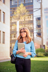 Blond haired middle aged business woman holding laptop in her hand and walking in the city