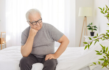 Sad senior man in grief sits in on bed in comfortable bedroom, deep in thought and thinking during...